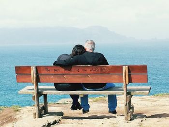 Rear view of men sitting on bench by sea against sky