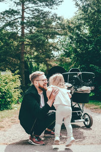 Happy man crouching while holding daughter by baby carriage on footpath at park