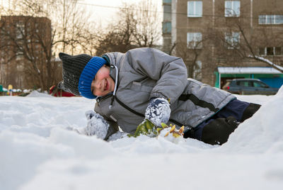 Smiling caucasian boy in green jacket and blue knit cap is lying on the snow.