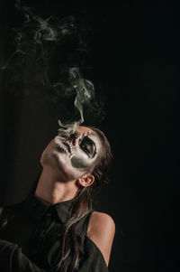 Portrait of young woman with face paint smoking against black background