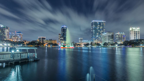 Illuminated city at waterfront against cloudy sky