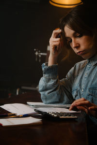 Side view of focused female worker using calculator to count income while sitting at counter with documents during work in cafe