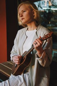 Thoughtful young woman playing guitar against window