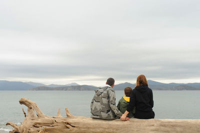 Family of three sits back old tree  on sandy beach and admires the blue clear sea and mountains