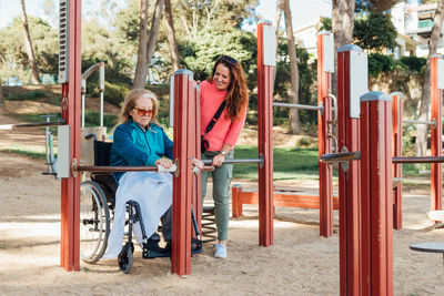 Content adult woman helping senior mother in wheelchair doing exercises on sports ground during rehabilitation session