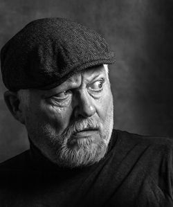 An elderly man with a beard and a cap looks to the side in shock. the picture is a  portrait.