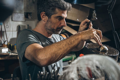 Side view of mature male artisan using metal punch and hammer to carve ornament on silver lid while sitting at table in professional workshop