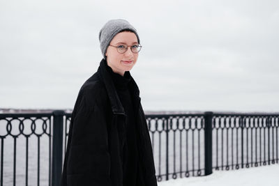 Smiling young woman with short hair in hat, eyeglasses, black coat looking at camera