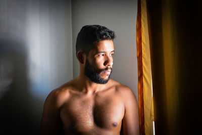Portrait of shirtless man looking away while standing against wall