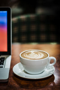 Close-up of cappuccino by laptop on wooden table