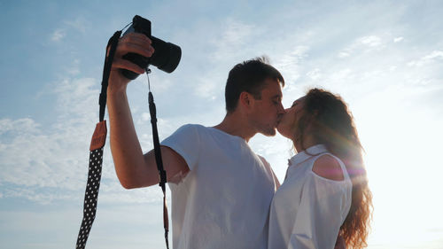 Young couple kissing on mouth against sky during sunny day