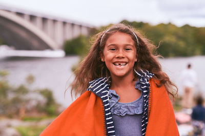 Portrait of cheerful girl with orange blanket standing in park during picnic