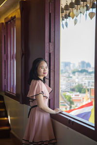  asian woman in a pink dress, she is looking at a camera standing next to a window 