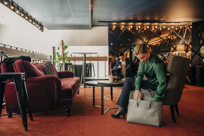 Mature businesswoman searching in bag while sitting on chair at hotel lounge