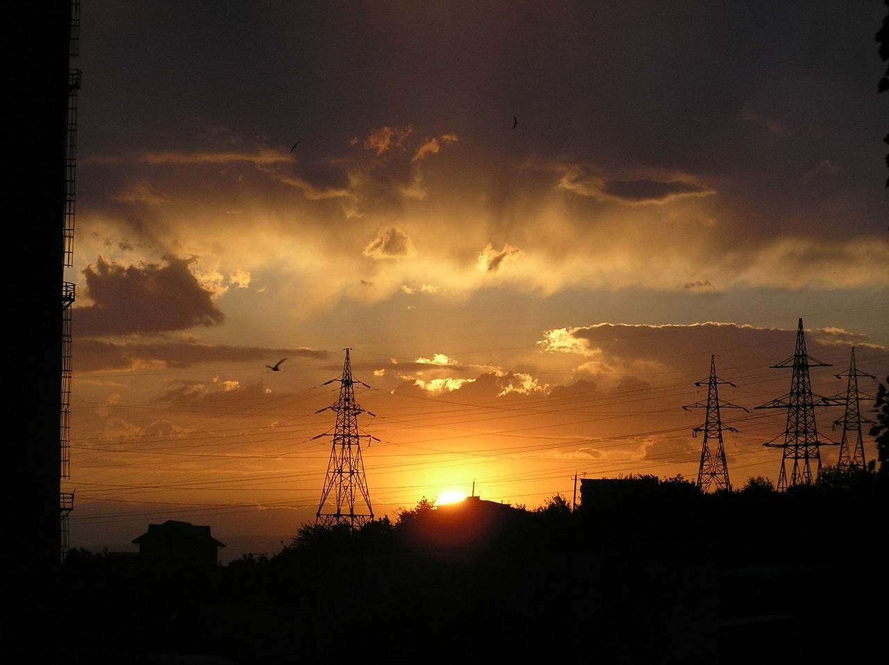 sunset, silhouette, power line, electricity pylon, electricity, power supply, sun, sky, fuel and power generation, orange color, connection, cable, cloud - sky, low angle view, building exterior, technology, sunlight, built structure, beauty in nature, nature