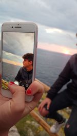 Cropped hand photographing teenage boy against sea during sunset