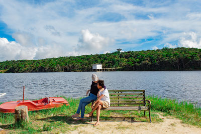 Couple sitting on bench by lake against sky