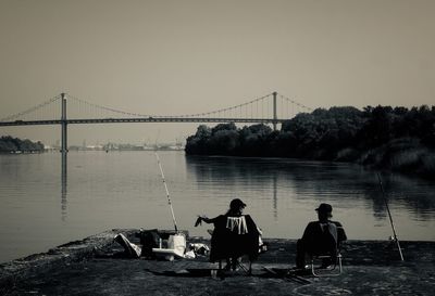 Rear view of friends fishing in river against bridge during sunset