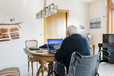 Back view of senior man in wheelchair using laptop at home