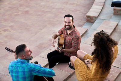 Content male musicians playing songs on acoustic guitars and looking at each other while sitting on steps with cheerful woman on street