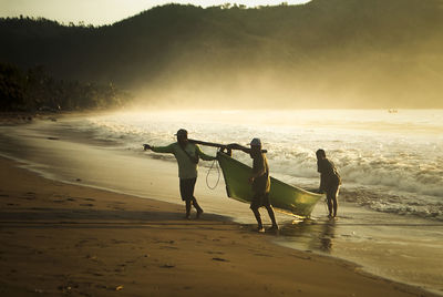 Fishermen carrying boat at beach against sky during sunset