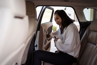 Crop positive ethnic female passenger in formal wear with smartphone entering in car on backseat