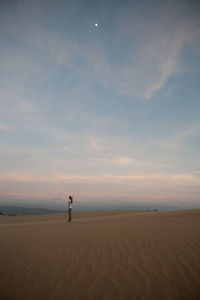 Side view of woman standing on sandy beach against sky during sunset