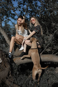 Female friends with dog sitting on log against trees in forest