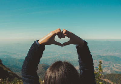 Rear view of young woman making heart shape on mountain against blue sky