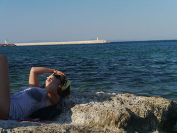 Woman sitting on retaining wall by sea against clear sky