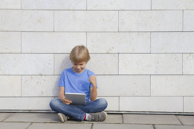 Full length of boy using digital tablet while sitting against wall