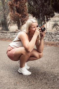 Side view of young woman photographing while crouching on road
