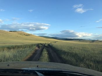Dirt road amidst field against sky seen through car windshield