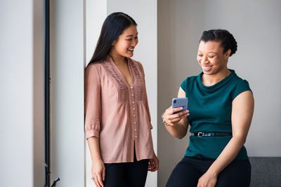 Happy female colleagues looking at smart phone against wall in office