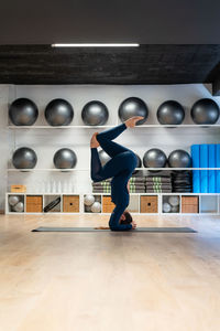 Full body side view of fit female in sportswear doing supported headstand variation pose while practicing yoga in fitness studio