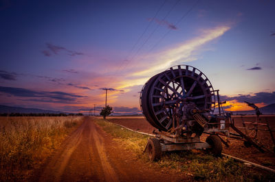 Agricultural machinery on dirt road against sky during sunset