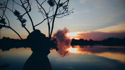 Silhouette man emitting smoke by lake against sky during sunset