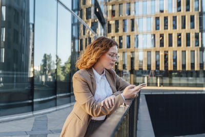 Young redhead businesswoman using mobile phone leaning on railing