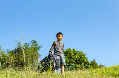 Full length of boy holding backpack while walking on grass against clear sky