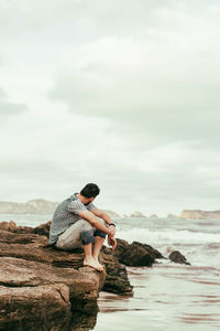 Side view of mid adult man sitting on rock at beach against cloudy sky