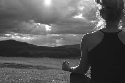 Grayscale image of a lady who is meditating on a field faced to the sun behind stormy sky