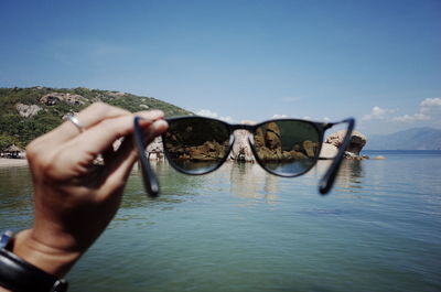 Midsection of person holding sunglasses against sea against sky