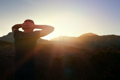 Rear view of man standing with hands behind head on field against clear sky during sunset