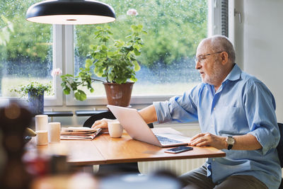 Senior man with laptop reaching for newspaper at table
