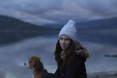 Portrait of smiling woman with beagle sitting by lake against sky during winter