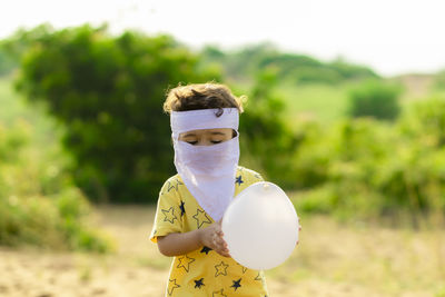 A little cute baby wearing a home made white full face mask and holding a white balloon in his hands