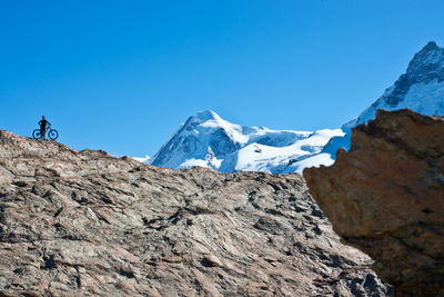 Low angle view of man with bicycle standing on rocky mountain against clear blue sky