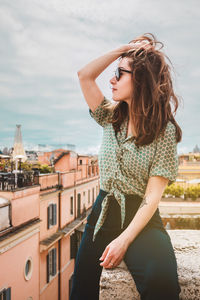 A young smiling woman in sunglasses sits on a rooftop with a panoramic view of rome, italy