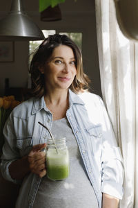 Smiling pregnant woman with glass of green smoothie standing at home