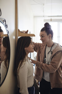 Smiling school nurse measuring height of female student standing by wall